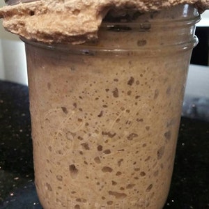 May include: A clear glass jar containing a light brown, bubbly mixture, possibly a sourdough starter. The jar has a silver lid and a brown, textured substance around the rim. The text "Anything Healthy" is visible on the lid.