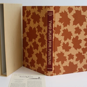 May include: A brown book with a gold and brown patterned cover. The title "Two Plays for Puritans" by George Bernard Shaw is printed on the spine. The book is in a beige box.