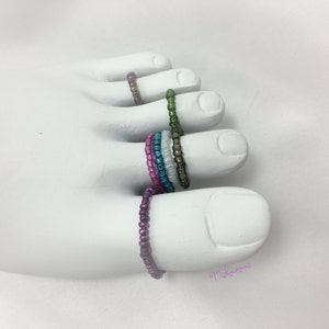 May include: A set of five colorful beaded toe rings. The rings are made of small, iridescent beads in shades of pink, blue, green, and white. The rings are stacked on a white mannequin foot.