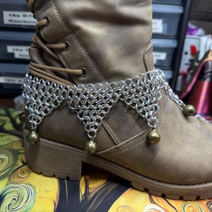 May include: A silver chainmail anklet with dangling brass-colored bells is wrapped around a brown leather boot. The chainmail has a geometric design. The boot has laces and a thick sole. The background is blurred, showing a work area.