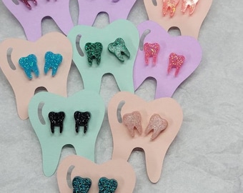 May include: Assortment of glitter tooth earrings displayed on tooth-shaped cards. The earrings are in various colors, including pink, purple, teal, black, and green.