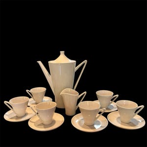 May include: A cream-coloured porcelain tea set, comprising a tall coffee pot, creamer, sugar bowl, and six teacups with saucers. The set features a minimalist design with clean lines and a glossy finish, ideal for serving tea or coffee.