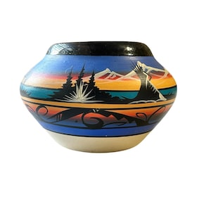 Puede incluir: Una olla de cerámica pintada a mano con borde negro y base color crema. La olla presenta un diseño de paisaje con montañas, árboles y una puesta de sol en tonos azules, naranjas y negros. La olla tiene aproximadamente 15 cm de diámetro.