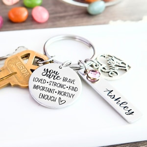May include: Silver keychain with a round charm that reads "You are brave loved strong kind important worthy enough" with a small heart at the end. The keychain also has a small silver charm with the word "awesome" and a pink rhinestone charm. A silver tag with the name "Ashley" is attached to the keychain.