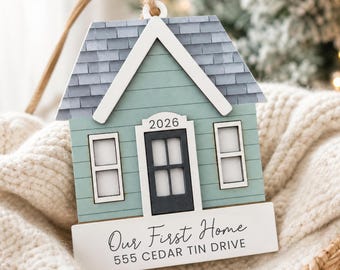 May include: A house-shaped ornament with a light green facade, a dark gray roof, and a black door. The text 2026 is above the door. A white base features the text Our First Home and 555 CEDAR TIN DRIVE. A tan cord is attached for hanging.