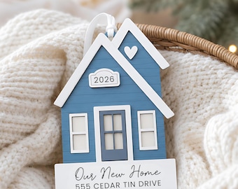 May include: A blue house-shaped ornament with white trim, featuring a small heart, a plaque with the year 2026, and a white base printed with the text Our New Home and 555 CEDAR TIN DRIVE. The ornament includes a white ribbon for hanging.