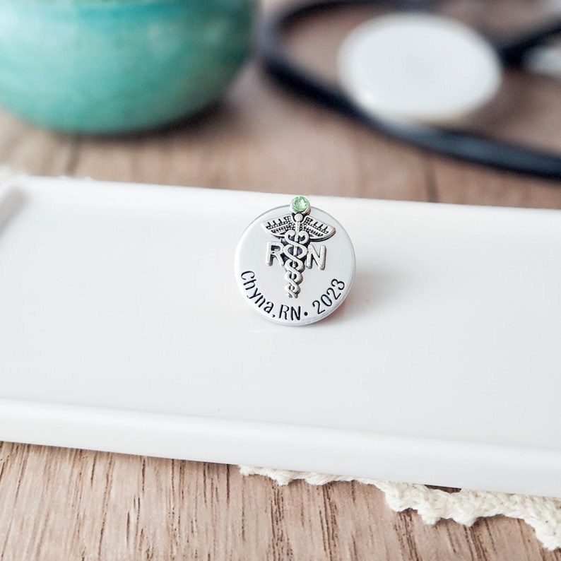 Puede incluir: Pin de plata con una piedra preciosa verde, un s&iacute;mbolo de caduceo y el texto "RN" y "Chyna. RN. 2023".