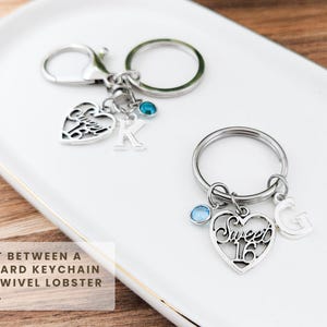 May include: Two silver keychains with heart-shaped charms. One keychain has a charm with the letter "K" and a blue gem. The other keychain has a charm that says "Sweet 16" and the letter "G" with a blue gem. The keychains are on a white tray.