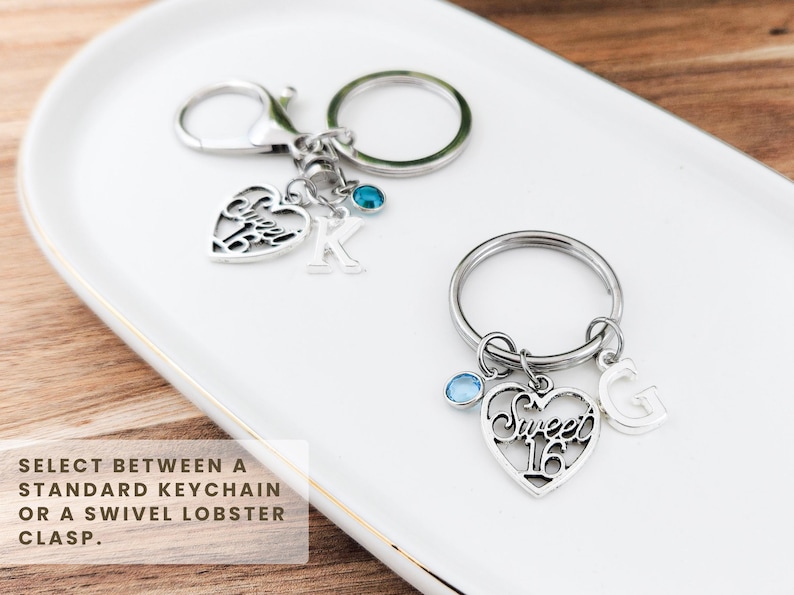 May include: Two silver keychains with heart-shaped charms. One keychain has a charm with the letter "K" and a blue gem. The other keychain has a charm that says "Sweet 16" and the letter "G" with a blue gem. The keychains are on a white tray.