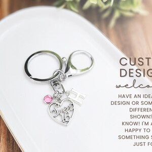 May include: Silver keychain with a heart-shaped charm that says "Sweet 16" and a pink gem. Includes a letter "H" charm. The text "CUSTOM DESIGNS welcome" is also visible.