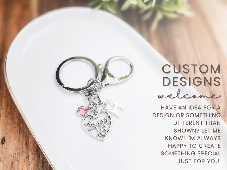 May include: Silver keychain with a heart-shaped charm that says "Sweet 16" and a pink gem. Includes a letter "H" charm. The text "CUSTOM DESIGNS welcome" is also visible.