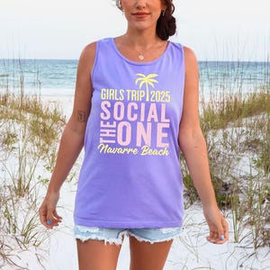 May include: A light purple sleeveless tank top featuring a yellow palm tree icon and text that reads GIRLS TRIP 2025, THE SOCIAL ONE, and Navarre Beach in a mix of yellow and light pink lettering printed on the front of the garment.