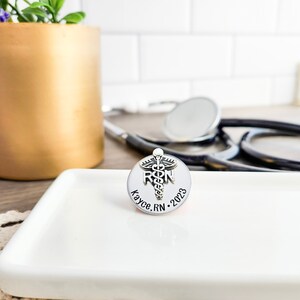 Puede incluir: Pin de plata con un s&iacute;mbolo de caduceo y las letras "RN" en una base redonda. La base est&aacute; grabada con el texto "Kayce, RN 2023".