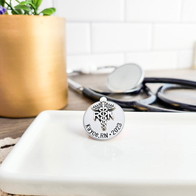 Puede incluir: Pin de plata con un s&iacute;mbolo de caduceo y las letras "RN" en una base redonda. La base est&aacute; grabada con el texto "Kayce, RN 2023".
