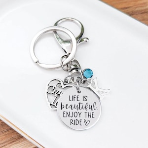 May include: Silver-toned keychain with multiple charms. A round charm reads "LIFE IS beautiful. ENJOY THE RIDE." A heart-shaped charm and a small key charm are also attached. A blue gem adds a pop of color.