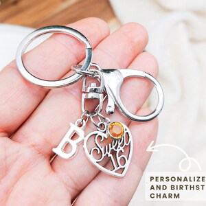 May include: Silver keychain with a heart-shaped charm that says "Sweet 16". Includes a letter "B" charm and a small orange gemstone. The text on the image says "Personalize Initial and Birthstone Charm".