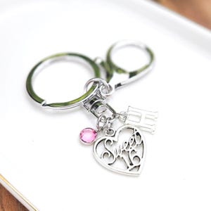 May include: Silver-toned keychain with a heart-shaped charm that says "Sweet 16". The charm has a pink gemstone and the letter "H". The keychain is on a white surface.