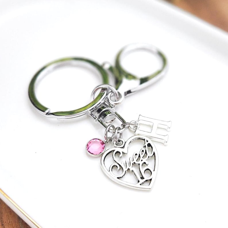 May include: Silver-toned keychain with a heart-shaped charm that says "Sweet 16". The charm has a pink gemstone and the letter "H". The keychain is on a white surface.