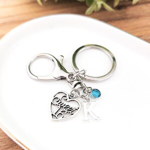 May include: Silver-toned keychain with a heart-shaped charm that reads "Sweet 16". The keychain also features a letter "K" charm and a small blue gemstone. The keychain is on a white, oval-shaped plate.