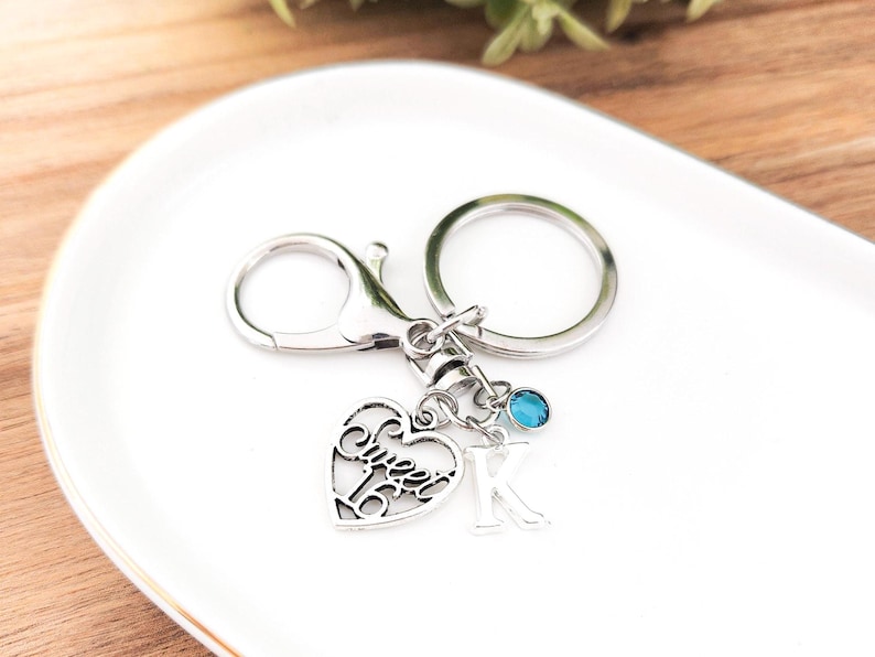 May include: Silver-toned keychain with a heart-shaped charm that reads "Sweet 16". The keychain also features a letter "K" charm and a small blue gemstone. The keychain is on a white, oval-shaped plate.