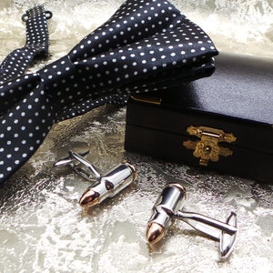 Może przedstawiać: Czarny krawat muszkowy w białe kropki, para srebrnych spinek do mankietów w kształcie pocisku i czarne pudełko ze złotym zamkiem.