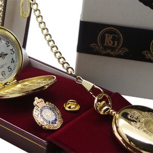 May include: A gold pocket watch with a white face and black Roman numeral hour markers. The watch is in a red velvet box with a gold chain and a gold pin with a crest. The box is labelled "KG Company, Exclusive Gold Gifts."