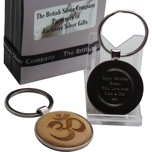 Puede incluir: Un llavero de plata con un dije redondo de madera con un símbolo Om grabado. El llavero está empaquetado en una caja blanca con cinta negra y el texto "The British Silver Company Purveyors of Exclusive Silver Gifts".