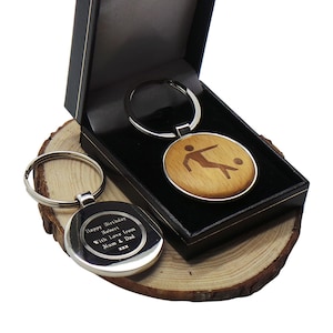 May include: A silver key chain with a round wooden charm featuring a silhouette of a soccer player kicking a ball. The back of the charm is engraved with the text "Happy Birthday Robert With Love from Mum & Dad xxx".