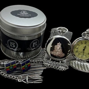 Könnte beinhalten: Ein silbernes Taschenuhren-Set mit Kette, Manschettenknöpfen und einer Blechdose. Die Taschenuhr hat ein silbernes Zifferblatt mit schwarzen römischen Ziffern und ein silbernes Gehäuse mit einem gravierten Design. Die Manschettenknöpfe sind silberfarben mit einem blauen und gelben Emaille-Design. Die Blechdose ist silberfarben mit einem schwarzen Etikett, auf dem "British Silver Company" steht.