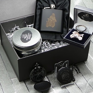 May include: A black gift box filled with a silver hip flask, a silver motorcycle keychain, a silver pocket watch, and a tin of tea. The hip flask has a motorcycle design engraved on it. The keychain and pocket watch also have motorcycle designs. The tin of tea has a label that says "British Silver Company".