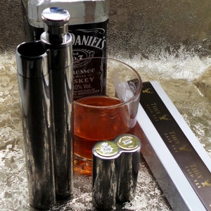 Puede incluir: Naturaleza muerta con una botella de whisky, un vaso de líquido ámbar y varios frascos plateados. También hay un pequeño frasco de doble cámara con tapas en forma de moneda. Una caja con el texto "THEY SAID YES" es visible.