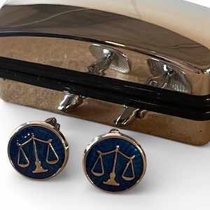 Puede incluir: Un par de gemelos con fondo de esmalte azul marino y diseño dorado de la balanza de la justicia. Los gemelos redondos tienen un borde plateado. Una caja plateada está en el fondo.