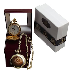 May include: A gold pocket watch with a white face and black Roman numeral hour markers. The watch is in a red velvet box with a gold chain. The box is closed and the watch is visible through the glass lid. The watch has the words "Lest We Forget" engraved on the back.