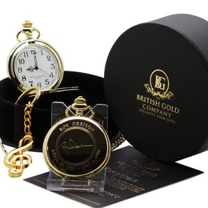 May include: A gold pocket watch with a white face and black Roman numeral hour markers. The watch is engraved with "Roy Orbison" and the dates "April 25, 1936 - Dec 6, 1988". The watch is in a black velvet box with a gold chain and a gold music note charm.
