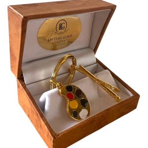 Puede incluir: Un llavero de paleta de artista de color dorado con manchas de pintura roja, negra, amarilla y blanca. El llavero incluye dos pinceles dorados y se presenta en una caja marrón con una etiqueta dorada que dice "The British Gold Company".