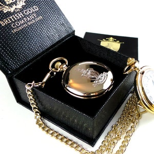 May include: A gold pocket watch with a chain in a black box. The watch has a white face with black Roman numeral hour markers and a gold case with an engraved design. The box is labeled "The British Gold Company Exclusive Gold Gifts".
