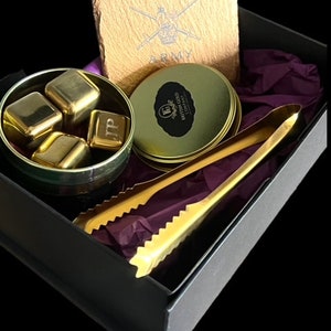 May include: A black gift box with a purple lining contains a set of gold metal whiskey stones, a gold metal tong, a round gold tin with a black label that reads "British Gold Whisky Liqueur", and a square brown stone coaster with the words "Army" printed on it.