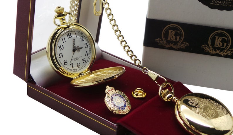 May include: A gold pocket watch with a white face and black Roman numeral hour markers. The watch is in a red velvet box with a gold chain and a gold pin with a crest. The box is labeled "KG Company, Exclusive Gold Gifts."