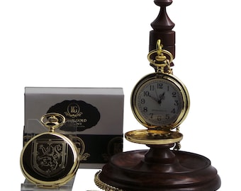 Puede incluir: Un reloj de bolsillo dorado con una esfera blanca y marcadores de hora romanos negros. El reloj está en un soporte de madera con una cadena dorada. El reloj está en una caja con el texto "British Gold Company" en ella.