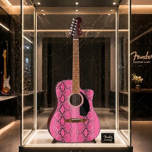 May include: Acoustic guitar with a pink snakeskin pattern on display in a glass case. The guitar has a dark brown neck and headstock, with the Fender logo visible. The case is lit with spotlights, and the Fender Custom Shop logo is visible.