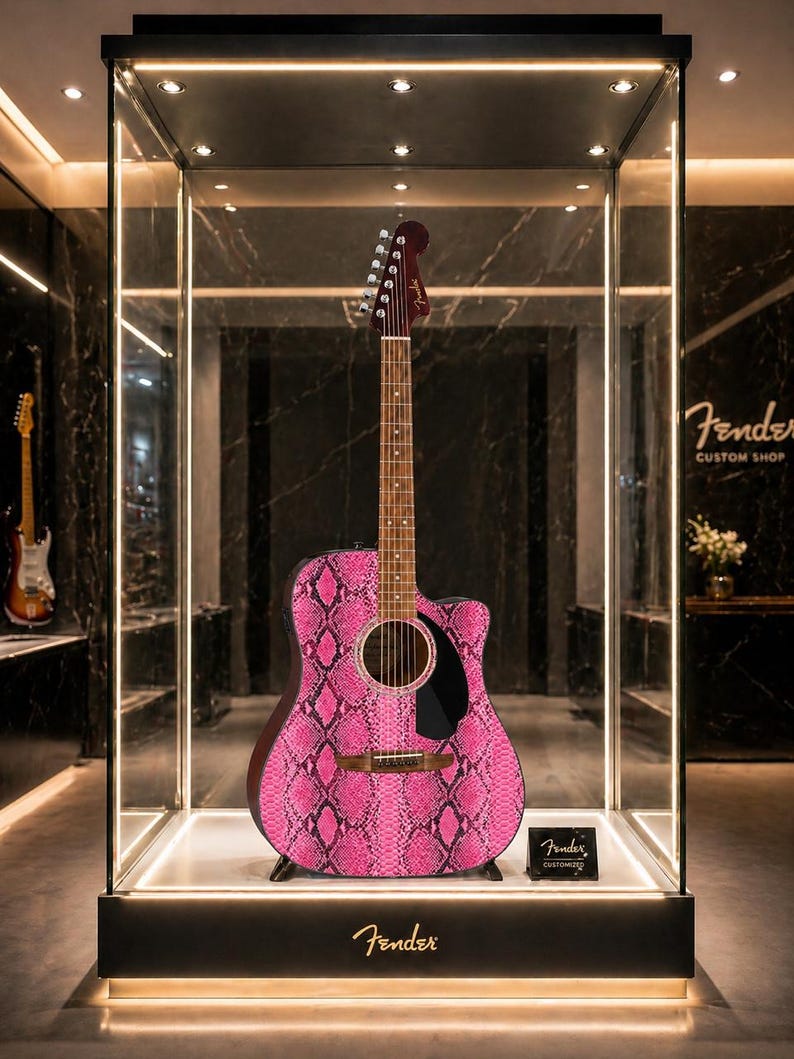 May include: Acoustic guitar with a pink snakeskin pattern on display in a glass case. The guitar has a dark brown neck and headstock, with the Fender logo visible. The case is lit with spotlights, and the Fender Custom Shop logo is visible.