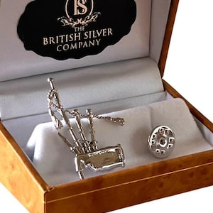 Puede incluir: Un pin de gaita plateado y su cierre se muestran dentro de una caja de madera con interior blanco. La caja contiene una etiqueta negra con el texto "THE BRITISH SILVER COMPANY".