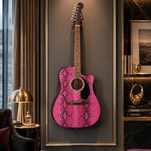 May include: Acoustic guitar with a vibrant pink snakeskin pattern. The guitar is mounted on a dark gray wall, framed with gold trim. The headstock is brown, and the sound hole is black. The guitar is a Fender.