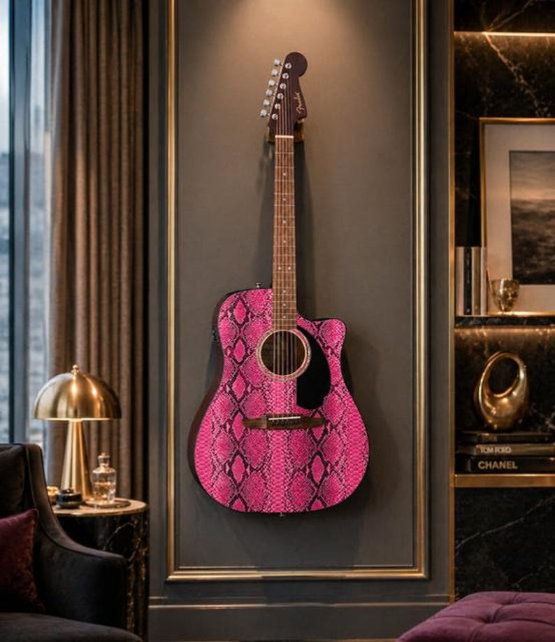 May include: Acoustic guitar with a vibrant pink snakeskin pattern. The guitar is mounted on a dark gray wall, framed with gold trim. The headstock is brown, and the sound hole is black. The guitar is a Fender.