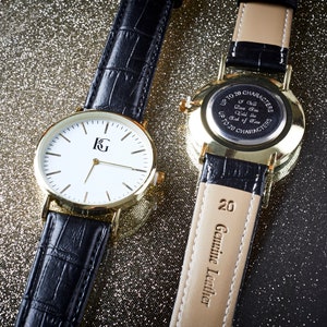 Op de afbeelding: Twee goudkleurige polshorloges met zwarte leren banden. Het horloge aan de linkerkant heeft een witte wijzerplaat met zwarte wijzers en een goudkleurig 'KG'-logo. Het horloge aan de rechterkant heeft een gouden achterkant met een inscriptie die luidt 'Maximaal 20 tekens - E&eacute;n zal er altijd zijn - Tot het einde der tijden - Maximaal 20 tekens'. De band van het rechterhorloge heeft '20 Echt Leder' erop gedrukt.