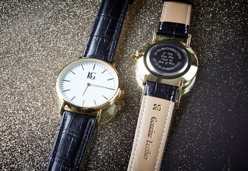 Op de afbeelding: Twee goudkleurige polshorloges met zwarte leren banden. Het horloge aan de linkerkant heeft een witte wijzerplaat met zwarte wijzers en een goudkleurig 'KG'-logo. Het horloge aan de rechterkant heeft een gouden achterkant met een inscriptie die luidt 'Maximaal 20 tekens - E&eacute;n zal er altijd zijn - Tot het einde der tijden - Maximaal 20 tekens'. De band van het rechterhorloge heeft '20 Echt Leder' erop gedrukt.