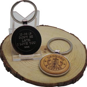 May include: Two keychains with different designs. The first keychain is silver with a black round tag that says "12-06-18 Don't Be Late! I Love You XXX". The second keychain is silver with a round wooden tag that says "Cannabis & Coffee" with a stylized image of a mermaid.