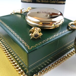 May include: A gold pocket watch with a white face and black Roman numeral hour markers. The watch is in a green box with a gold chain. The box has a gold Masonic symbol on the top.