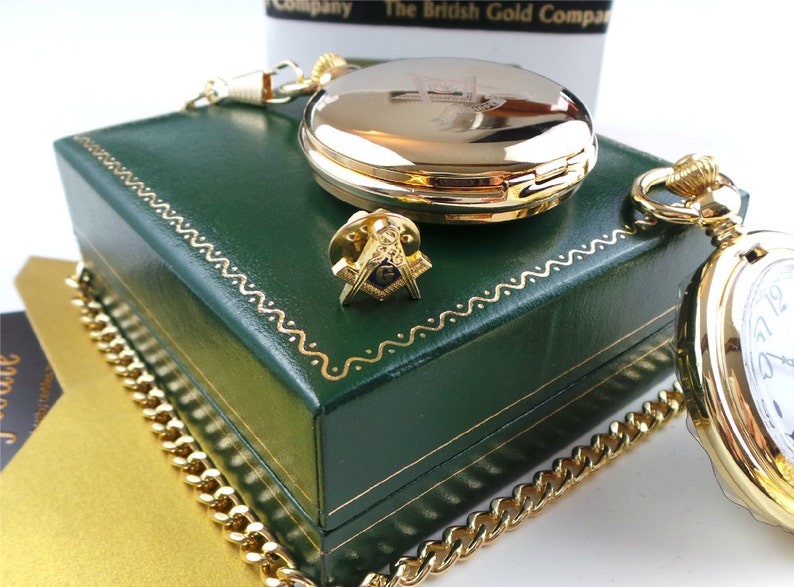 May include: A gold pocket watch with a white face and black Roman numeral hour markers. The watch is in a green box with a gold chain. The box has a gold Masonic symbol on the top.