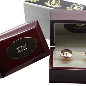 May include: Gold-toned cufflinks in the shape of a car's gear shift, presented in a dark wood box. The box is lined with cream-colored fabric. The cufflinks are displayed in a dark wood box with the words "British Gold Company" on the inside.